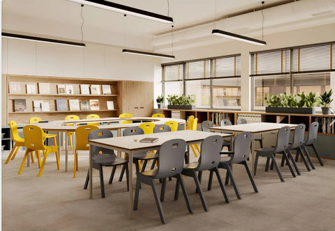 A modern classroom or study space with long rectangular tables and chairs. The foreground shows gray chairs around light wood tables, while the background includes more tables with yellow chairs, built-in bookshelves, large windows with blinds, and potted plants.